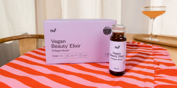 Vegan Beauty Elixir Verpackung und Fläschen auf einem Tisch mit gestreifter Tischdecke und Glas im Hintergrund